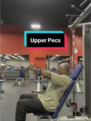 A man is seated on an incline bench within a gym setting, performing an exercise. The man is wearing a tan t-shirt and dark pants. Overlaid on the image is a rectangular text box, with the words "Upper Pecs."