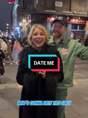 A woman and a man are standing outdoors on a city street at night. The woman is in the foreground, smiling at the viewer, with a black coat and fur trim. Behind her to the right, the man smiles. A sign in the background says "Nell of Soho". Overlaid on the image is a graphic with text.