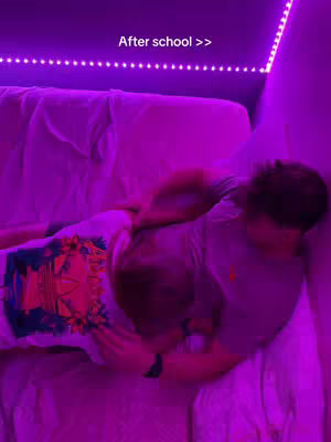 Two young men are lying on a bed, partially obscured by each other, in a room illuminated with a purple hue. The bed linens appear rumpled, implying a relaxed setting. Above, a string of purple LED lights casts a soft glow on the scene. The text 'After school >>' is visible above the bed, suggesting the video's theme or timing.