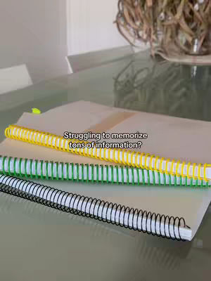 The image shows three spiral notebooks resting on a light-colored surface, likely a desk or table. The notebooks are stacked; the top one is yellow, the middle one is green, and the bottom one is black. A hand-written text overlay reads, "struggling to memorize tons of information?"