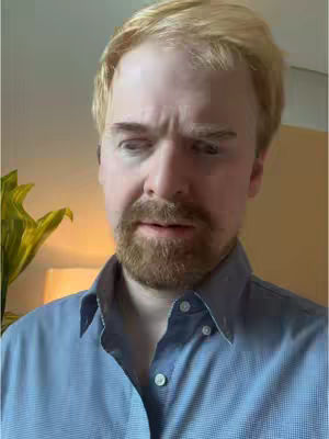 A man with fair skin, light brown hair, and a styled, light brown beard looks down with a concerned expression. He wears a blue button-down shirt. The background is blurred with soft lighting.