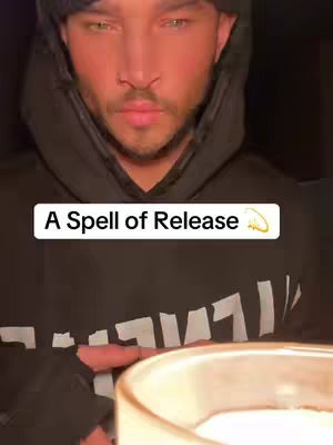 A man with a beard wearing a black hoodie is captured in a close-up shot, set against a dark background. A text overlay reading 'A Spell of Release' is positioned on the image. He appears to be holding something in front of him, possibly a candle, with his gaze focused downward. The lighting seems dim, casting shadows that draw attention to the man's face.