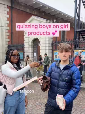 The cover image shows two young people, a Black woman and a white man, outdoors. The woman on the left is holding out a tampon, labeled with the text "tampon," towards the man on the right, who looks confused. A text overlay reads "quizzing boys on girl products," with two pink heart emojis. The background shows a brick building and other people.