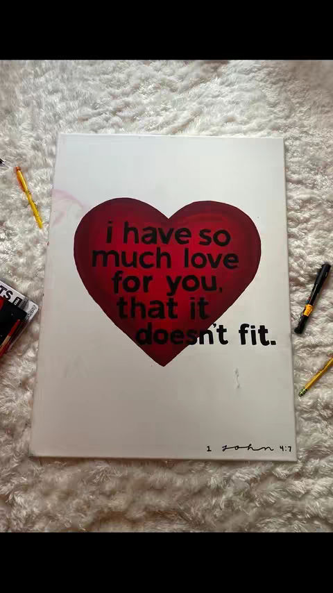 A white canvas with a large, red heart shape dominating the center. Inside the heart, black text states "i have so much love for you. that it doesn't fit." Below the heart, in a smaller font, is the text "1 john 4:7". The canvas is resting on a white, textured surface, and various pens and pencils are visible at the edges of the canvas.