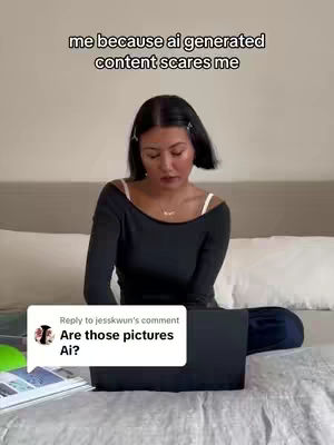 A woman sits on a bed with a laptop in front of her, looking at the screen. She is wearing a black top and the background is a bedroom setting.