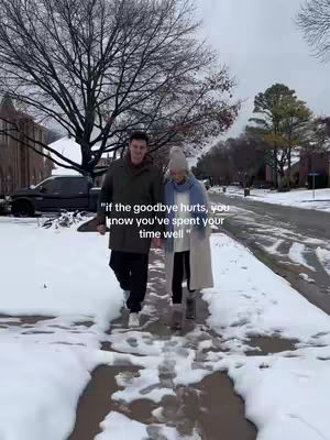 A couple walking on a snow-covered path. The man on the left is wearing a green coat and black pants. The woman on the right is wearing a white coat, a light-colored hat, and a scarf. The background features trees, some bare and some with a few leaves, and buildings along the street. The sky is overcast, suggesting it is a cold day. The path they are walking on is cleared of snow.