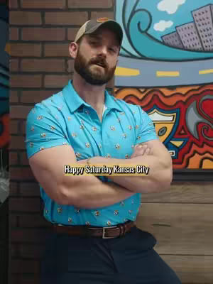 A man with a beard wearing a baseball cap and a patterned blue polo shirt stands with arms crossed. A vibrant mural is in the background.