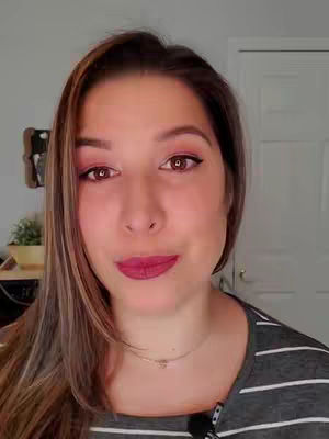 A medium shot of a woman's face, looking directly at the camera with a slightly amused expression. She has long, light brown hair with darker roots and is wearing pink eye shadow and lipstick. She has a small, delicate silver necklace. In the background is a wall with a door visible.