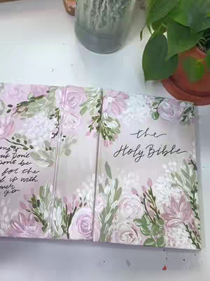 The cover image shows an artfully painted Bible with a soft, floral design. The Bible is open, displaying decorated pages with a light pink background and detailed floral motifs in pastel colors, including pink roses and green foliage. The words "the Holy Bible" are visible on the right side of the open book, written in an elegant script. The composition is bright and uses natural lighting, suggesting a handmade and personalized aesthetic.