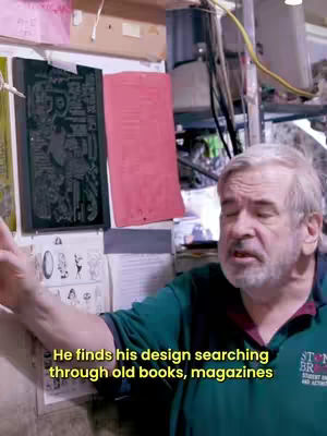 An older man with gray hair and a beard stands in what appears to be a workshop or store, gesturing towards a wall with various items displayed. A text overlay in yellow reveals additional information about the shot.