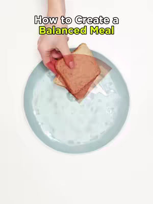 A top-down shot featuring a person holding a plate with a sandwich. The background is a light blue and white. The text overlay reads, "How to Create a Balanced Meal" with a green-yellow outline.