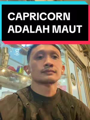 A man with short dark hair and a mustache is centered in the frame, looking at the camera with a neutral expression. Above him, the text 'CAPRICORN ADALAH MAUT' is displayed on a black background, bordered by cyan and red. The background is slightly blurred, suggesting a public setting. Behind the man, there's a display shelf with various items.