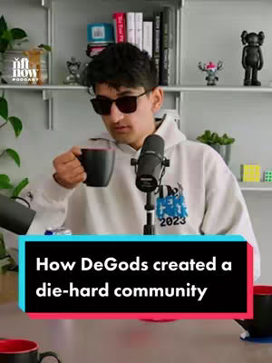 A man wearing sunglasses and a white hoodie holds a coffee mug while sitting in front of a microphone. There is a shelf with items in the background, and below the man, a banner with text.