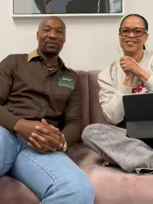 Two people are seated on a pink couch. A man wearing a brown shirt with a logo patch and blue jeans is on the left. A woman is seated on the right, wearing a light-colored sweater and glasses. A laptop is visible to the right of the woman.