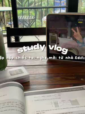 The cover image captures a study scene at a cafe. The main elements include an open book, a closed notebook, and a tablet displaying a video interface. A cup, likely containing a beverage, is visible in the upper left corner. The table is made of wood. The background suggests an outdoor setting with greenery visible through the window.