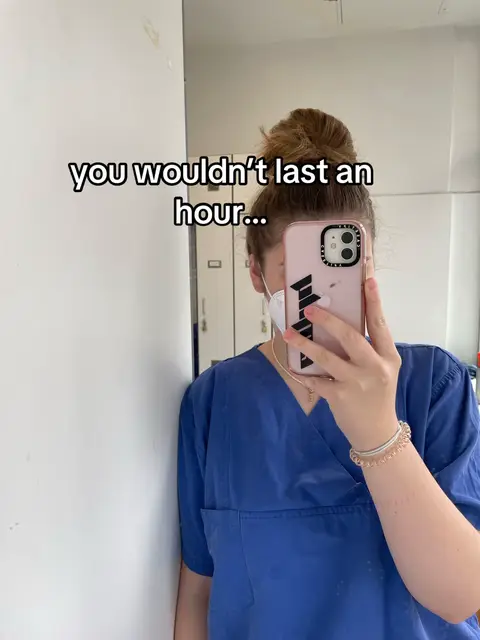 A woman wearing blue scrubs and a face mask is holding up her phone to take a selfie, obscuring most of her face. Text overlay reads 'you wouldn't last an hour...'. She's leaning against a white wall next to a mirror, suggesting a medical setting. Her hair is in a bun.