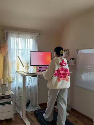 A person is standing on a treadmill desk in front of a computer monitor in a domestic setting. The person is wearing a star-adorned hoodie and sweatpants, with a single flower in their hair. The background includes a window with sheer curtains and a whiteboard. Soft, natural lighting fills the room.