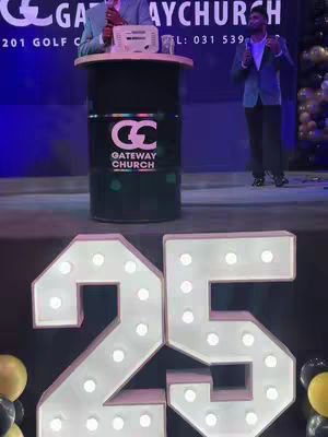 A stage setup with a large lit-up number '25', a podium with a church logo, and two men behind it. Balloons and blurred backgrounds add to the celebratory atmosphere.