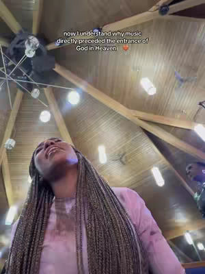 A woman with long, braided hair looks up toward a church ceiling with a complex wooden structure and hanging lights. The woman is wearing a pink top.