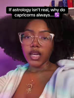 A woman with dark curly hair and glasses is facing the camera, with text overlayed. She is wearing a white and blue tie-dyed top and a necklace. The background is a blurry room with a purple light.