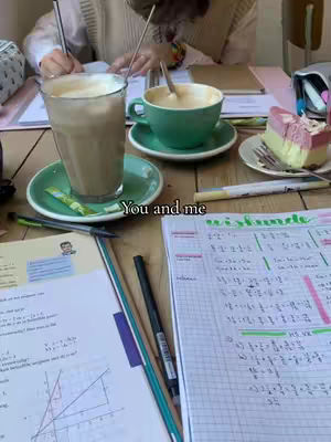 The image shows a table with study materials, two cups of coffee, and a piece of cake, suggesting a study session or date. A person is visible in the background with their hands on the table, writing. The color palette is muted with browns, greens, white, and pinks.