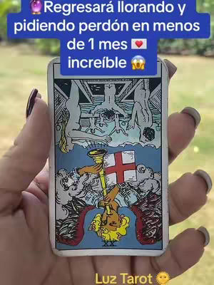 A hand holds a tarot card featuring a central illustration showing a figure, possibly an angel, holding a trumpet. Above, three figures are suspended. The card is on a white rectangle with a light blue background. A text overlay is placed at the top; the hand has long dark nails. The background appears to be a blurred outdoor scene.