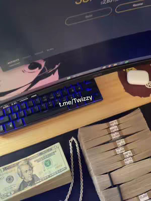 The image shows money, a keyboard, and a computer screen. Bundles of cash, a twenty-dollar bill, and a silver chain are on a dark wooden surface near a keyboard and monitor. The screen shows a website with a balance displayed. The keyboard has blue backlighting. The overall composition suggests wealth and online activity.