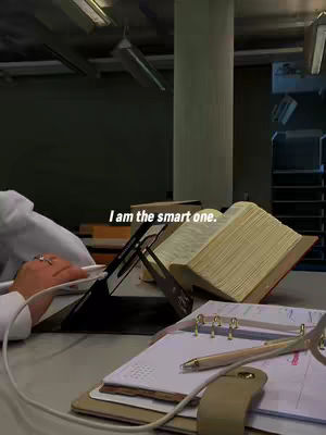 A workspace is shown with a person's hand resting on a touch pad. The foreground presents an open notebook with a pen, a tablet set with a cover, and an open book on a stand. The background includes a vertical pillar and some study lamps on the ceiling.