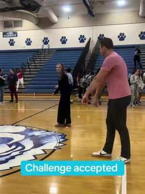 A man, wearing a pink shirt and black pants, stands at the edge of a basketball court, facing slightly to his left with arms outstretched, as a woman smiles and walks towards him. The court is bathed in the warm artificial light of a gymnasium, with a large logo at center court.