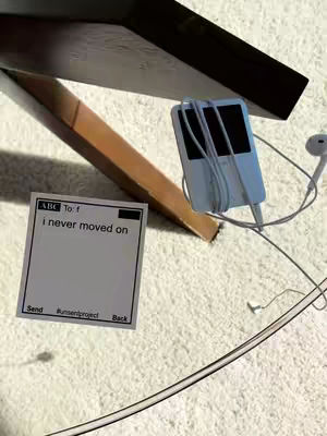 The image is a still life composition featuring a paper message, an iPod, and a partially visible wood object all placed on a shag carpet. The paper has text in a minimalistic style. An iPod with white earbuds is present. A angled wooden object is placed in the background, adding depth and perspective. The color palette consists of neutral tones—white, beige, brown and black.