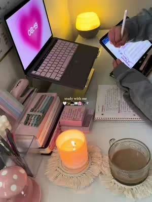 A desk setup with a laptop displaying a pink heart graphic, a person using a tablet, a candle, and a cup of coffee. The composition features a cozy and organized workspace.