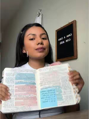 A young woman is holding an open Bible, which has notes and sticky notes inside. She is looking towards the camera, and behind her is a wall with a framed board that says 'ama a araq con 16:32' in mirror writing.