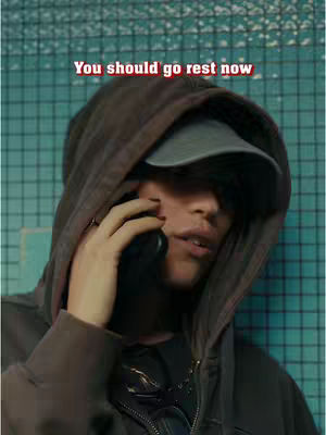 A person with a hood and cap is holding a phone to their ear, with text overlayed at the top that reads "You should go rest now". The background is slightly blurred and appears to be a wall of tiles.
