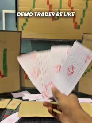 The cover image features the text 'DEMO TRADER BE LIKE' overlaid on a background that appears to be constructed of cardboard boxes, possibly representing a trading environment or a mock-up of one. The focus is primarily on a hand holding a fan of paper, which appears to have the outlines of bills with red stamps on them. The composition suggests a concept of demonstration, simulation, or a lighthearted take on trading activities. The lighting seems to be natural, with shadows indicating the depth and texture of the cardboard.