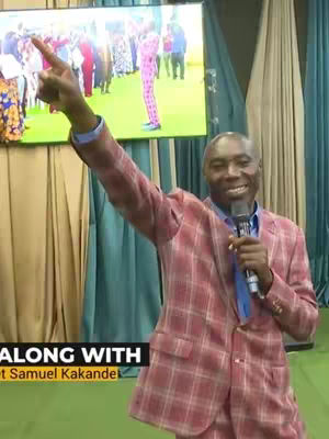 A man in a red plaid suit is holding a microphone and pointing with his right arm. Behind him is a large screen displaying people, and the scene is set in a studio with curtains.