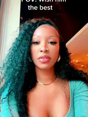 A selfie-style shot featuring a woman with long, curly teal hair. She has a delicate nose ring and is wearing a turquoise top. The background appears to be a restaurant or similar venue, with a window visible on the left and blurred out details.