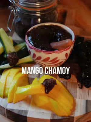 A colorful, appetizing food shot featuring a small, patterned bowl of dark sauce placed centrally. The bowl is surrounded by slices of bright yellow mango, green-skinned cucumber slices, and dark, textured pieces that appear to be dried fruit. A clear jar with a metal lid filled with a darker liquid is visible in the background. The text "MANGO CHAMOY" is spelled out in white, sans-serif font across the lower center.