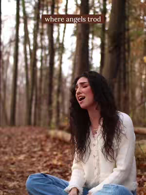A young woman with long dark hair is sitting outdoors. She is wearing a white sweater and blue jeans. She is looking upwards with her mouth open as if singing. Behind her are tall trees and a background of fall foliage. A text overlay displays the words 'where angels trod'.
