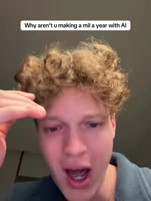 A close-up shot of a young man with curly blond hair. The man has a fair complexion and is looking towards the camera with an open mouth and wide eyes as if surprised. He has his right hand raised towards his head, as if to shade his eyes. The background is a blurred, dimly lit space with some darker patches, suggestive of an interior.