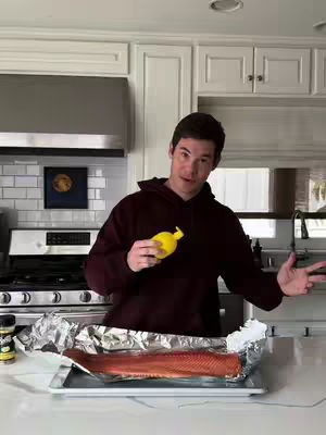 Adam Devine stands in a kitchen, holding a yellow lemon-shaped container above a preparation of salmon in front of him. He is smiling at the camera and gesturing with his other hand.