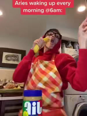 A person wearing glasses, a red hoodie, and an apron is shown in a kitchen setting. They are holding a banana towards their mouth as if they're about to take a bite. A jar of peanut butter is in the foreground. Text in a red box at the top says, 'Aries waking up every morning @6am'.