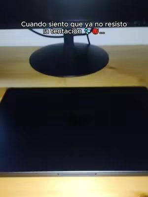 The image frames a desktop setup. In the foreground, there's a large, black screen of a laptop, and behind it, the stand of a monitor. Above the monitor stand is the monitor screen itself, displaying a light-colored background. The scene is lit, casting soft shadows, and the wooden desk is visible.