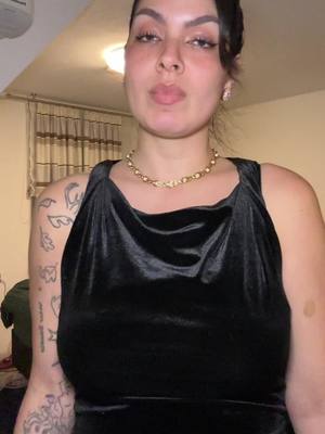 A woman is centered in a selfie-style shot, gazing upward with a slight pout. She wears a black dress with a cowl neckline and a gold necklace. Her dark hair is pulled back, and she has visible makeup and earrings. The background shows hints of a room's interior with curtains and a dim, warm light.