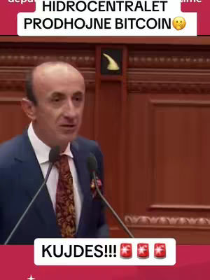 A man in a suit speaks behind a podium with microphones and a sign that says "HIDROCENTRALET PRODHOJNE BITCOIN" at the top. Below the man, a banner reads "KUJDES!!!" with three fire emojis.