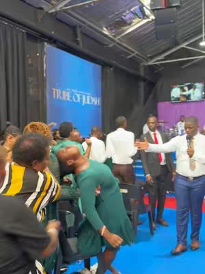 An interior shot of a large event with many people, featuring a woman in a green dress, with a teal backdrop. In the center, a woman in a green dress is leaning backward with her head tilted back, as if overcome with emotion. People are surrounding her, and a man in a suit is holding a microphone to the right. A blue screen is in the background with the text "TRIBE OF JUDAH".