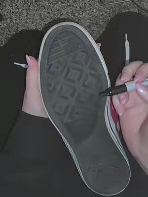 The image showcases the bottom of a white and red Converse shoe with writing on it. A hand holding a black marker is in the process of tracing the shoe sole pattern. The background is a blurry, dark surface.