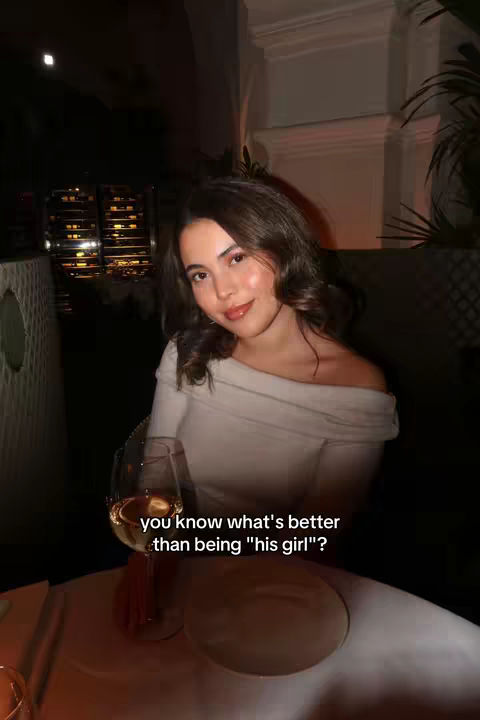 A woman sitting at a table in a restaurant, with a glass of wine in front of her. The background is dimly lit, suggesting a nighttime setting. She's looking at the camera with a slight smile.