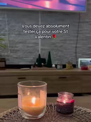 The cover image shows a cozy indoor setting, likely a living room. In the foreground, there are two candles, one inside a clear glass and another in a small jar. They are lit, creating warm, glowing light. The background has a wooden cabinet or shelf, and a screen displaying a purple, pink and red color gradient. The ambiance is intimate and relaxing, with the focus on the candles suggesting a romantic or celebratory theme.