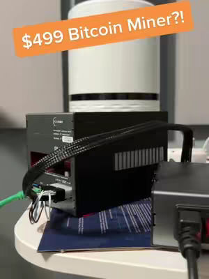The cover image displays a Bitcoin mining rig on a white surface. An orange banner with white text overlays the scene, reading '$499 Bitcoin Miner?!'. The rig includes a black box with power cables and an attached cylinder. The background is a blurry mix of gray and white.