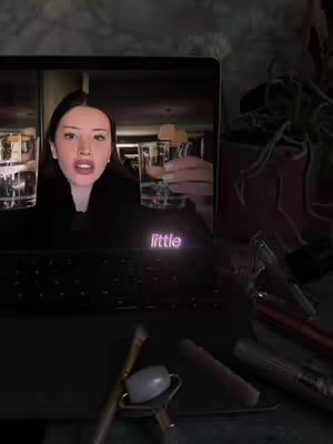 The cover image shows a laptop screen displaying a video of a woman holding two glasses of liquid, with the word "little" in pink neon font on the screen. The laptop is on a desk with makeup brushes and other items.